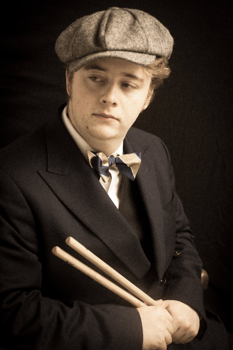 A young man looking to the left, holding a pair of drumsticks.